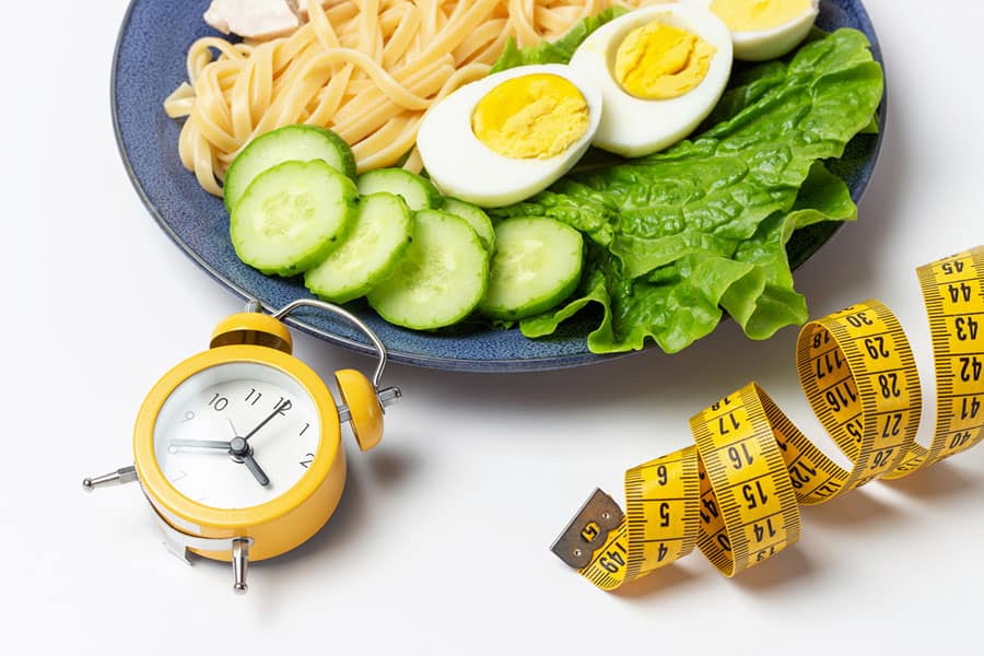 A plate full of healthy meal with an alarm clock and measuring tape lying on the side.