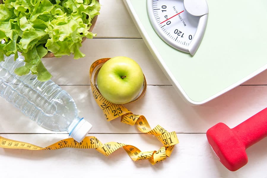 Components of healthy living: fresh fruits and veggies, water, exercise equipment and weight and measurement tools.