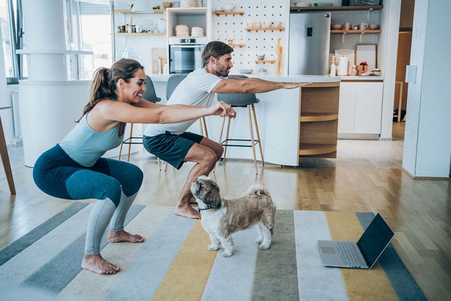 A couple doing basic squat exercises at home as their dog looks on