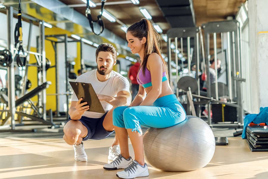 A trainer showing sample workout plan to a fitness enthusiast sitting on a swiss ball.