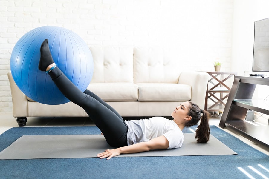 A woman lying on gray stretch mat and doing leg lifts with swiss ball.
