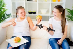 two teen girls are sitting on a couch. One is holding a burger in the hand with another burger placed on the lap and the other teen girl is holding a dumbbell.