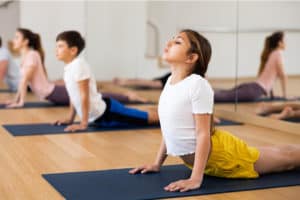A teenage girl is doing yoga on a yoga mat in a hall with her teenage peers