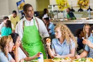Teens eating healthy food at a cafeteria while a cafeteria person is standing next to them