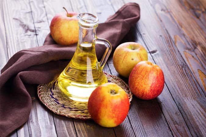 A glass jar of apple cider vinegar is placed on a table along with four apples.