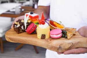 A serving board filled with macaroons, fruit tart, apple crumble, rainbow cake and a hazelnut brownie.