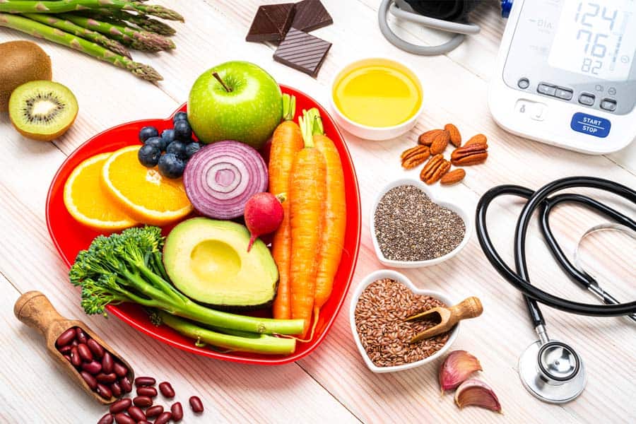 A plate full of vegan foods like carrot, avocado, onion and more is placed on a table near a blood pressure machine and other vegan foods.