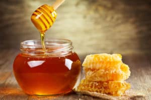 A person is holding a dipper wrapped in honey. Slices of honeycomb are placed near the honey jar.