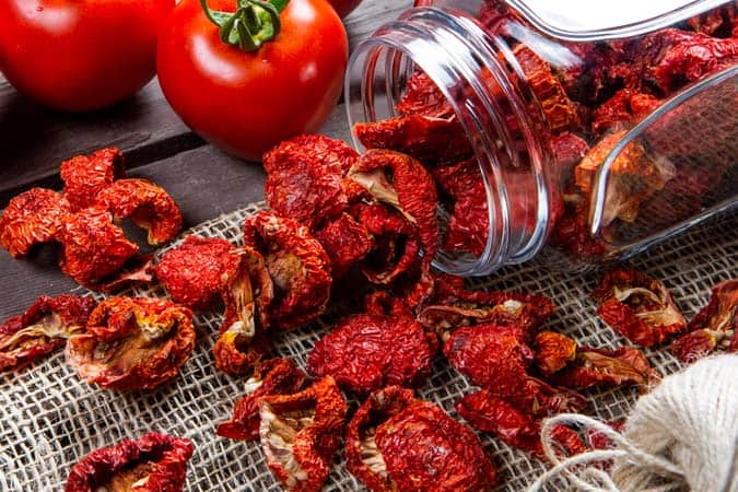 Sun-dried tomatoes are spilling out of a glass jar onto a table.