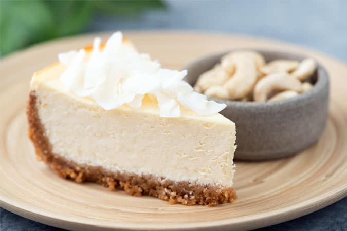 A slice of vegan cheesecake made with cashew cream served on a plate with a bowl of cashew nuts on the side.