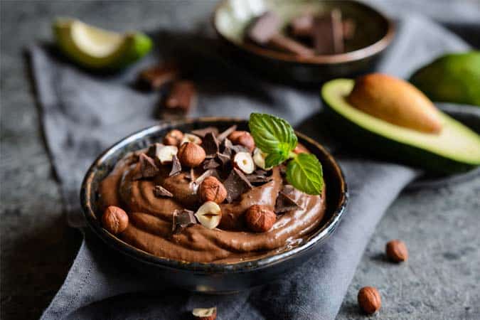 A small serving bowl with chocolate avocado mousse garnished with dark chocolate chunks, hazelnuts, and a sprig of mint.