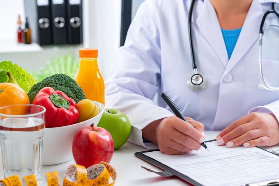 A dietician is seen filling a form. There is a bowl of fruits and vegetables along with a bottle of juice placed near her.