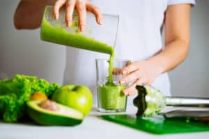 A female is seen pouring a vegan green colored smoothie from a jug to a glass. There are some fruits and a hand blender placed near her on the shelf.