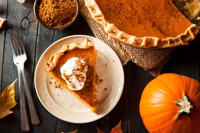 A slice of vegan pumpkin pie placed on a table. There is also a whole pumpkin, a big chunk of pumpkin pie, and a small bowl of spice on the table.