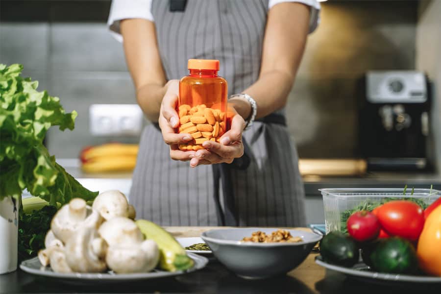 A female is holding a vegan supplement bottle while standing in a kitchen. Vegetables like mushroom, cucumber, tomatoes, lettuce are placed on the shelf in front of her.